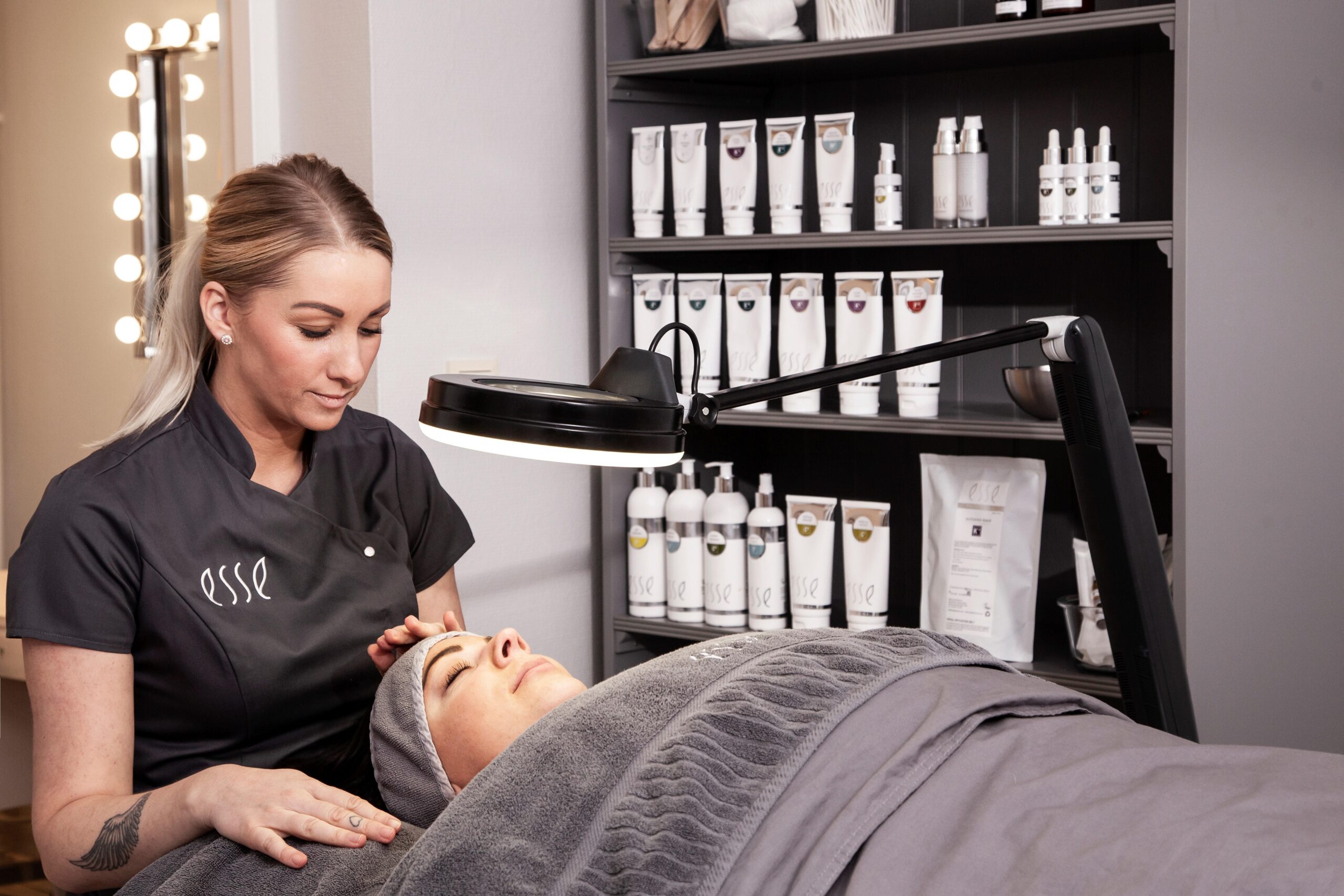 En hudpleiespesialist masserer forsiktig en klients ansikt i et spa-behandlingsrom. Kunden ligger på en behandlingsseng dekket av grå håndklær, og i bakgrunnen ser man hyller med ulike hudpleieprodukter.