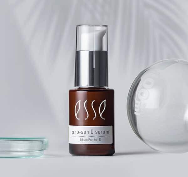 En brun glassflaske merket "Esse pro-sun D serum" med sølvfarget pumpehette står på en lys flate, ved siden av en klar, rund gjenstand og et glassfat, med en myk bladskygge i bakgrunnen.